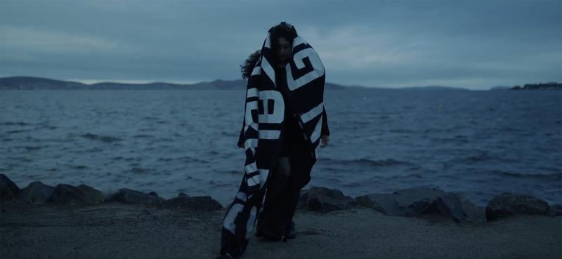 Baltimöre Charlót, standing on a dark beach wrapped in a black cloak with white symbols